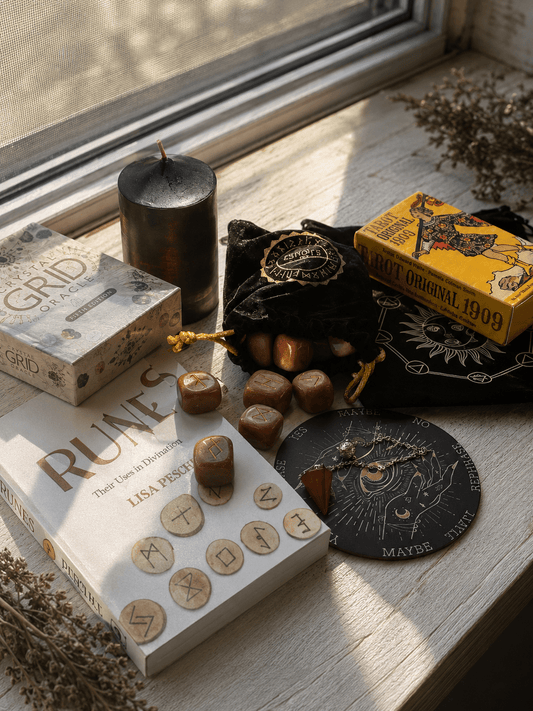 A styled flat-lay of a Wiccan/ pagan ritual kit on a sunlit windowsill. The kit includes a black candle, a black velvet pouch with gold trim, a yellow card deck box, a white book titled 'Runes', a gray box titled 'Grid', polished reddish-brown stones, a black circular tarot card deck, and dried botanical sprigs.