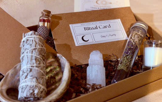 A curated ritual kit displayed inside a brown kraft box, featuring a sage smudge bundle in a shell dish, a white crystal ball, a glass jar of dried herbs, a small candle, and a card labeled 'Ritual Card Day 1: Purify'.