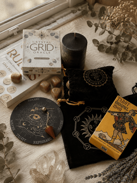 A styled flat-lay of a crystal grid oracle kit and tarot card deck on a cream fabric surface near a window. Visible items include a boxed Crystal Grid Oracle, a black candle, a black velvet pouch with gold printing, a round black oracle card deck, and a yellow tarot card box with illustrated artwork.