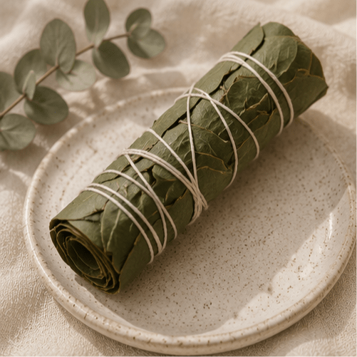 A rolled bundle of green leaves tied with white string, presented on a speckled ceramic plate with a eucalyptus branch in the background.