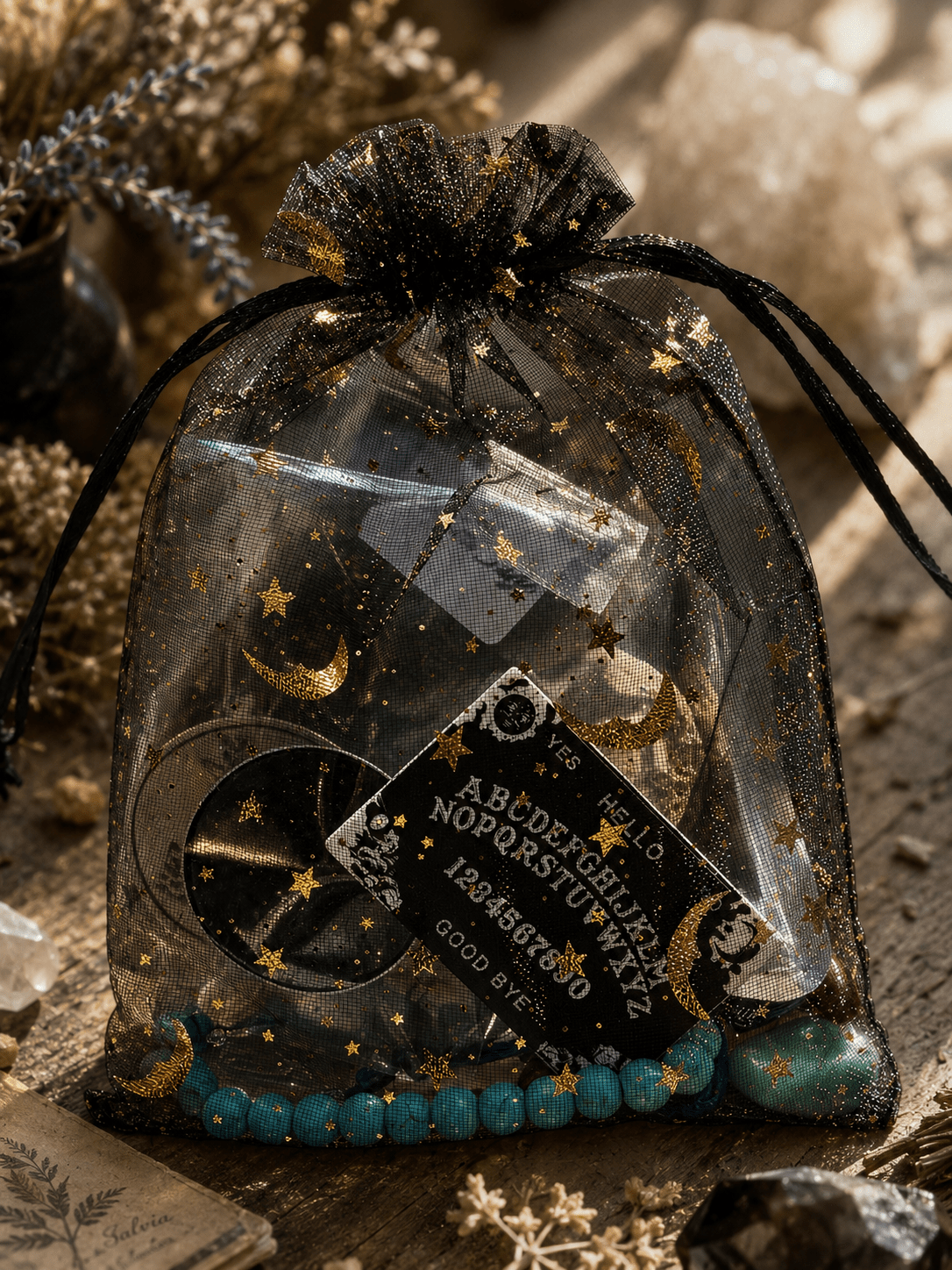 A black sheer organza drawstring pouch with gold star and moon prints, filled with a tarot card, a black coin, and turquoise beads, styled on a rustic wooden surface with dried flowers.
